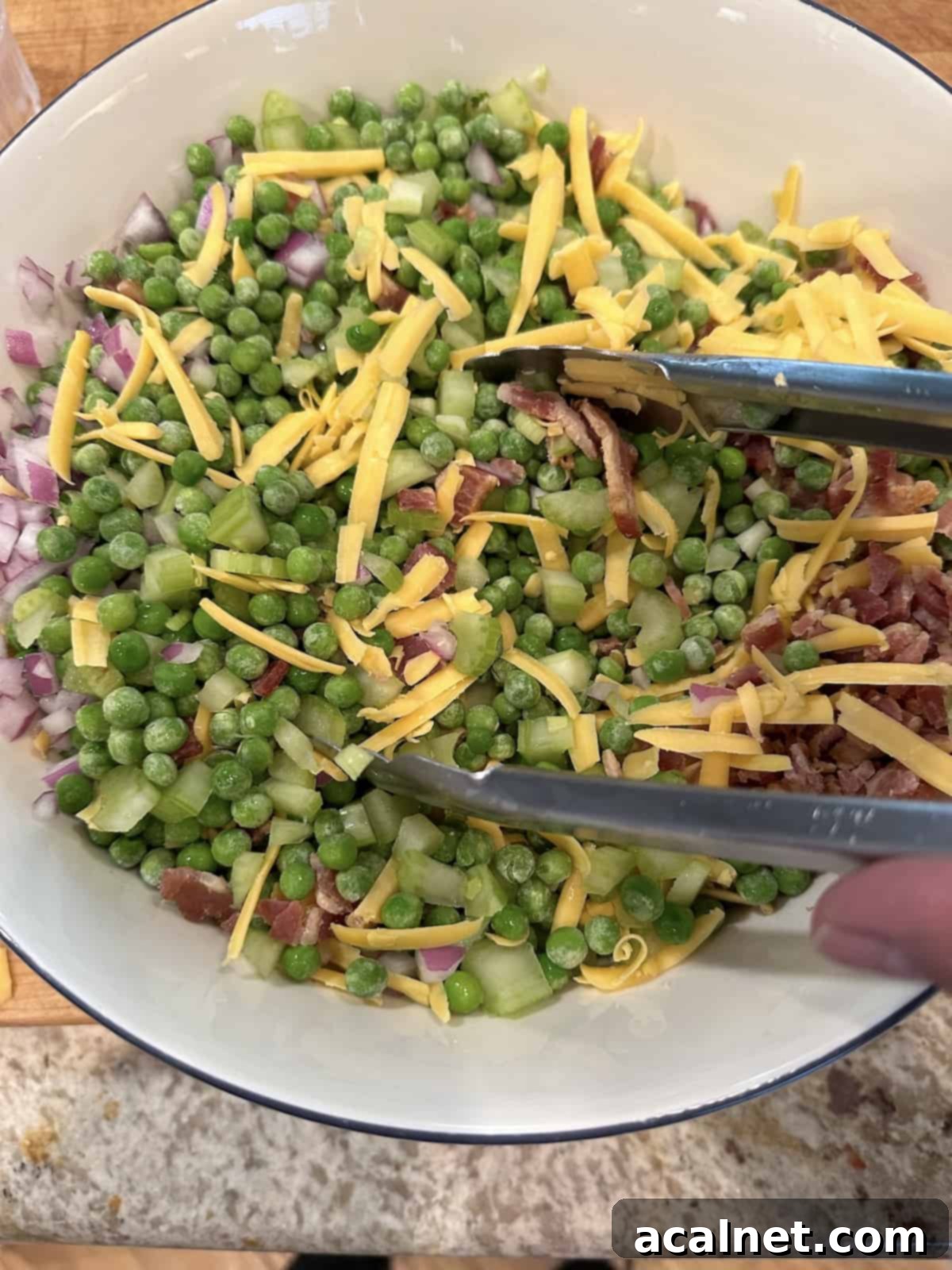A large glass bowl with thawed peas, shredded cheese, diced red onion, crispy bacon, and chopped celery being gently stirred with a pair of serving tongs.