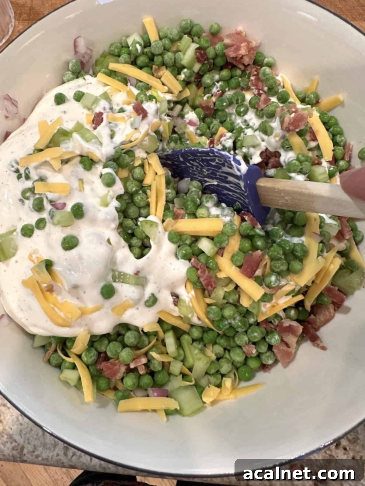 A large glass bowl of pea salad ingredients, with a generous dollop of the creamy white dressing placed on top, not yet fully mixed in.