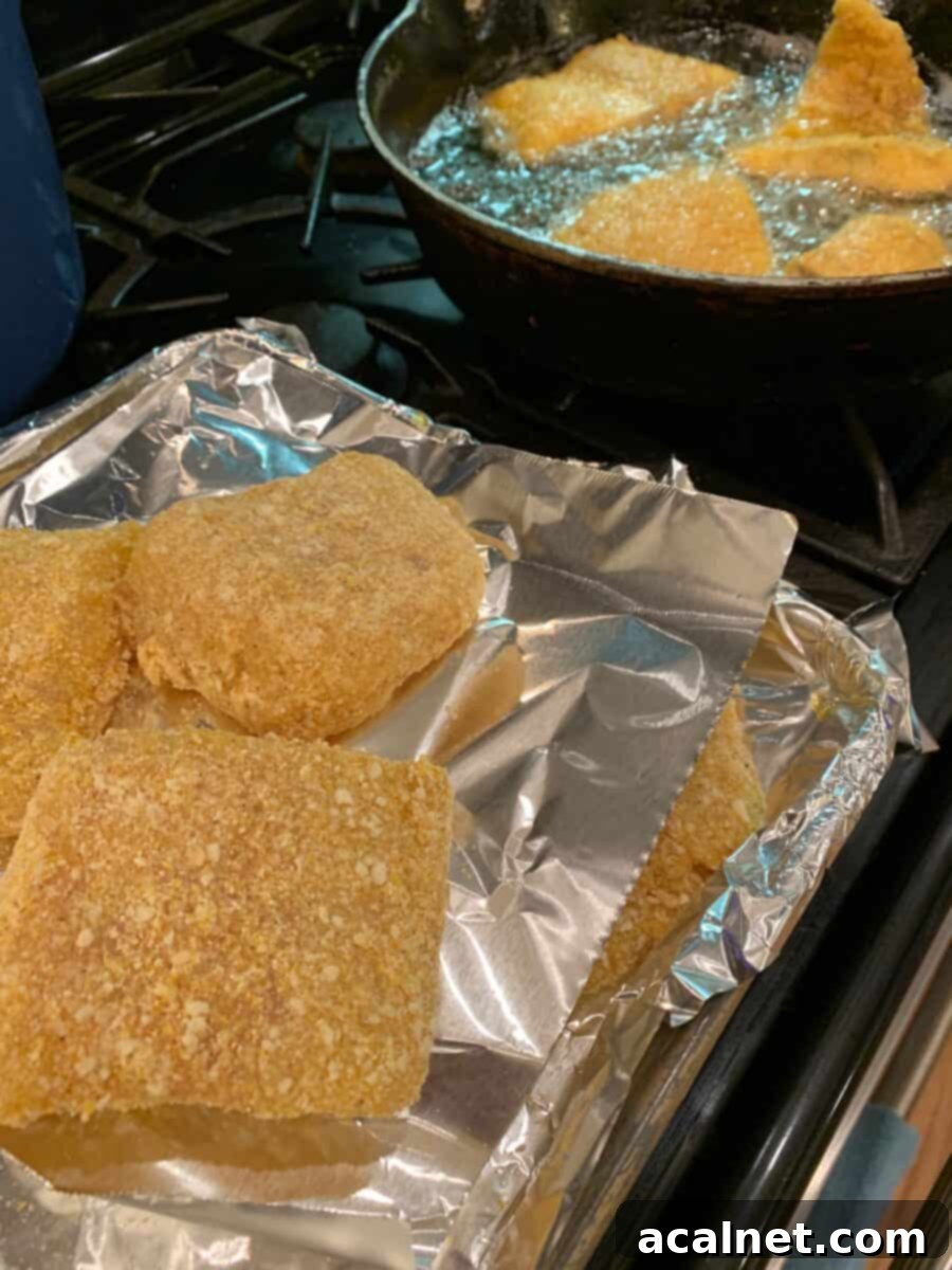 Breaded cod fillets waiting on foil next to a hot skillet where several pieces of fish are frying to a golden crisp.