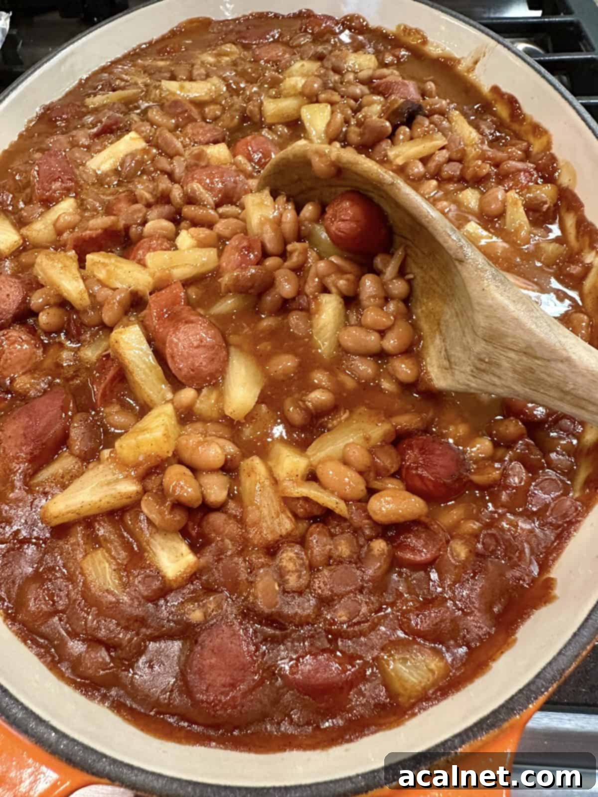 Franks, baked beans and pineapple slices in a baking dish with a wooden spoon, ready to be served.