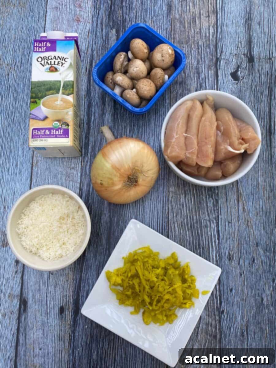Spicy Chicken Alfredo ingredients on a wooden block, displaying a carton of half and half, mushrooms, uncooked chicken, a sweet onion, grated parmesan cheese and a plate of pepperoncinis.