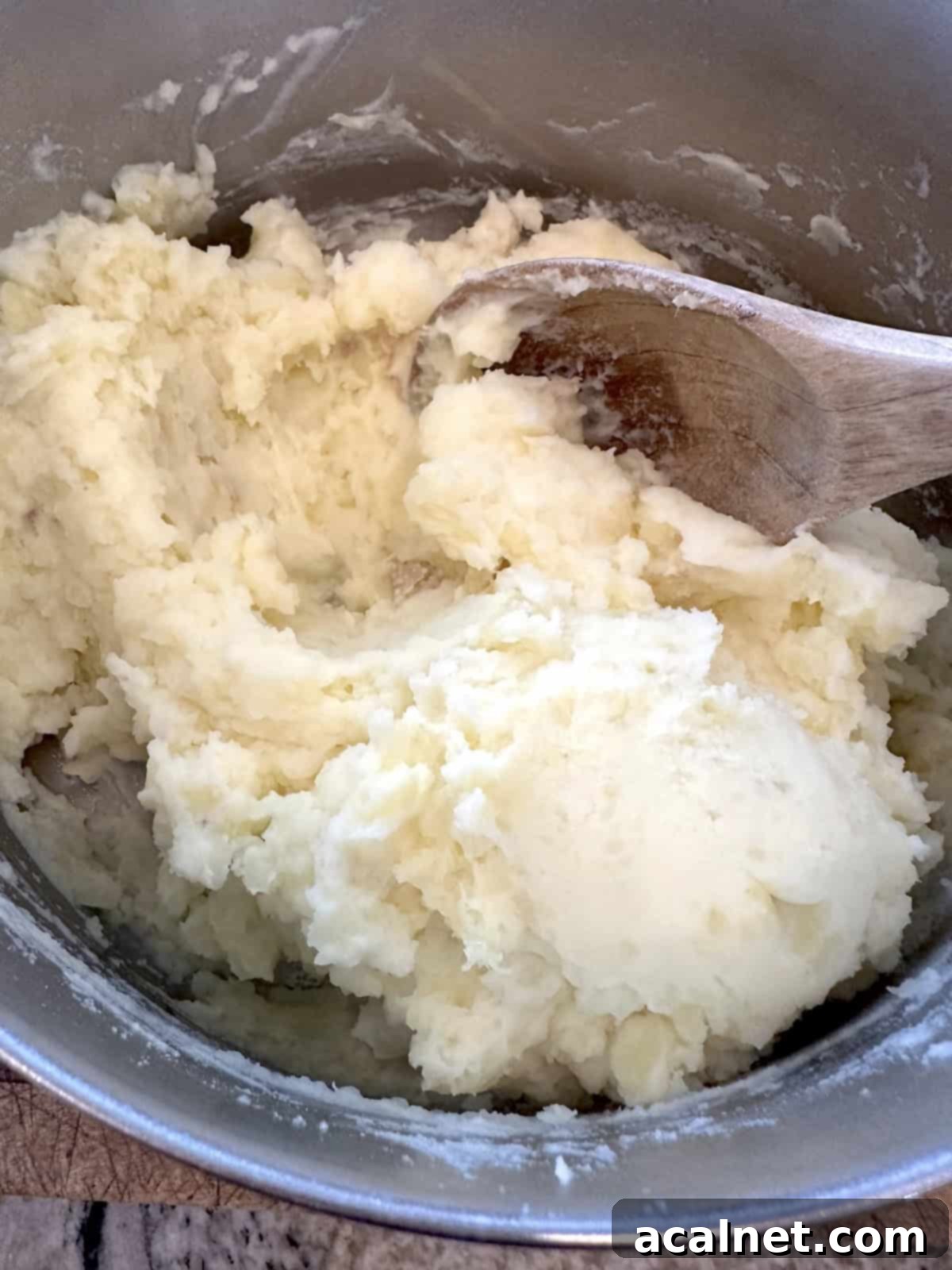 Mashed potatoes in a pan being stirred with a wooden spoon after liquid has been added.