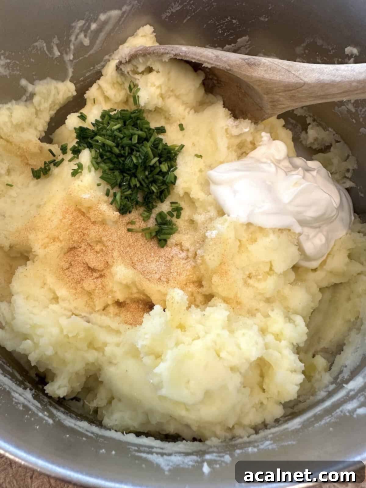 Mashed potatoes in a pot with sour cream, diced chives, and garlic powder being added.