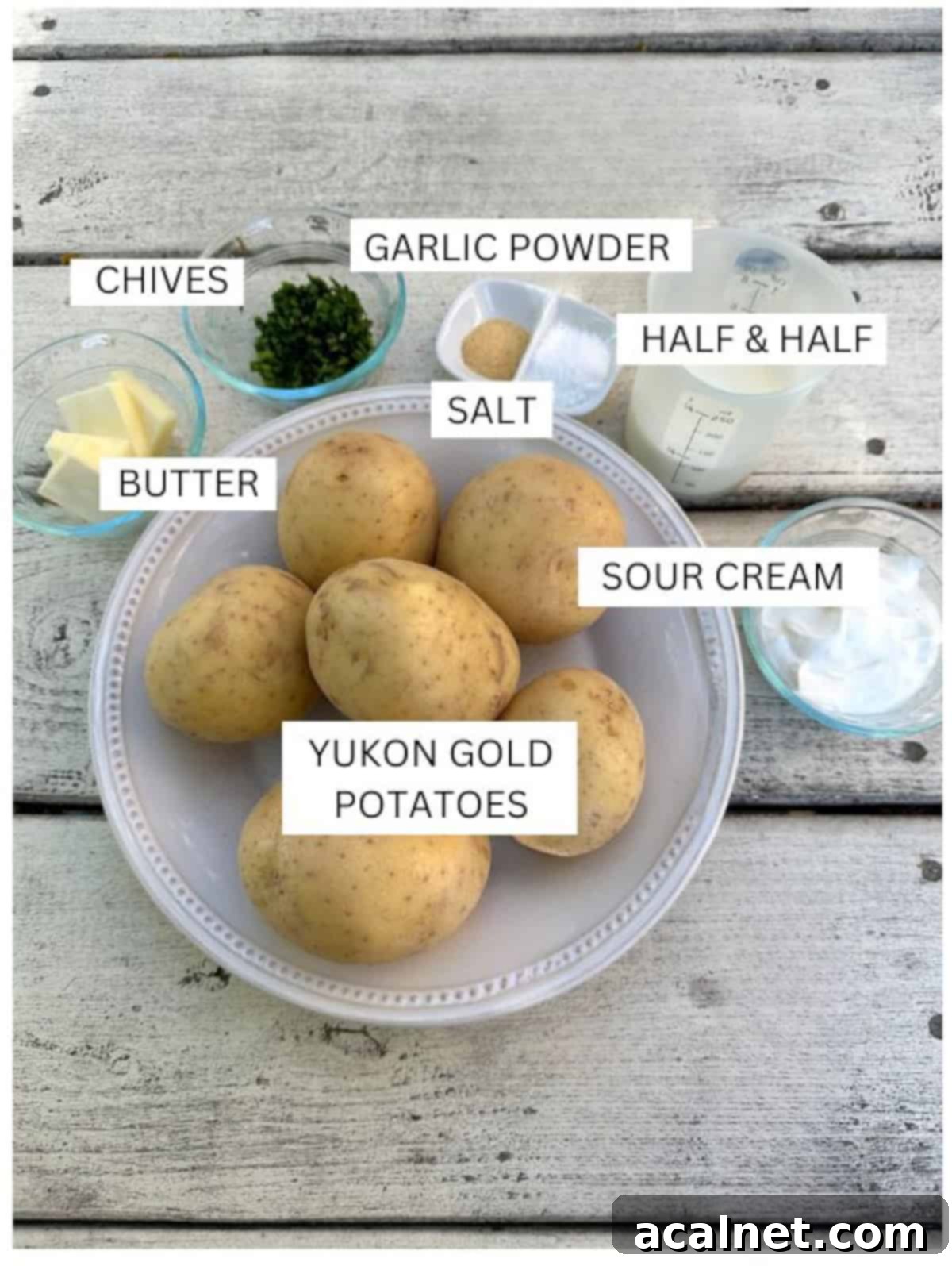 A selection of labeled ingredients including Yukon Gold potatoes, butter, milk, chives, and sour cream in small bowls on a wooden table.