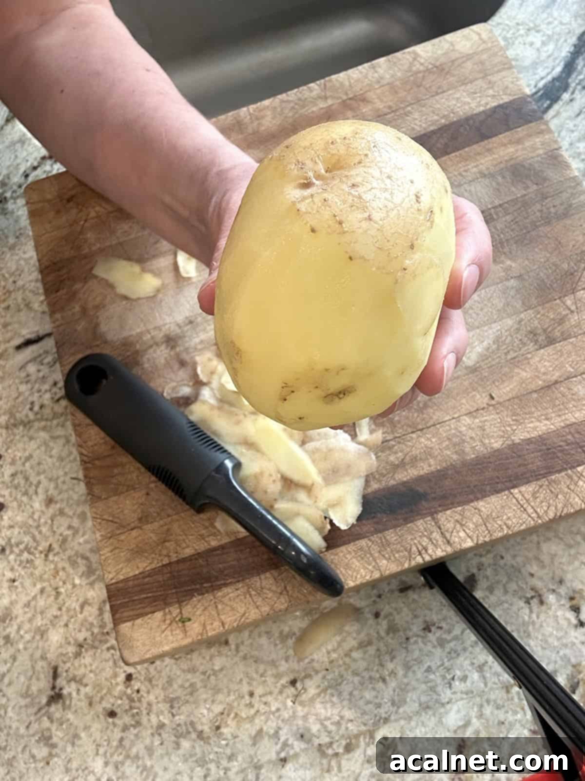 A single Yukon Gold potato, partially peeled with some skin remaining, on a cutting board.