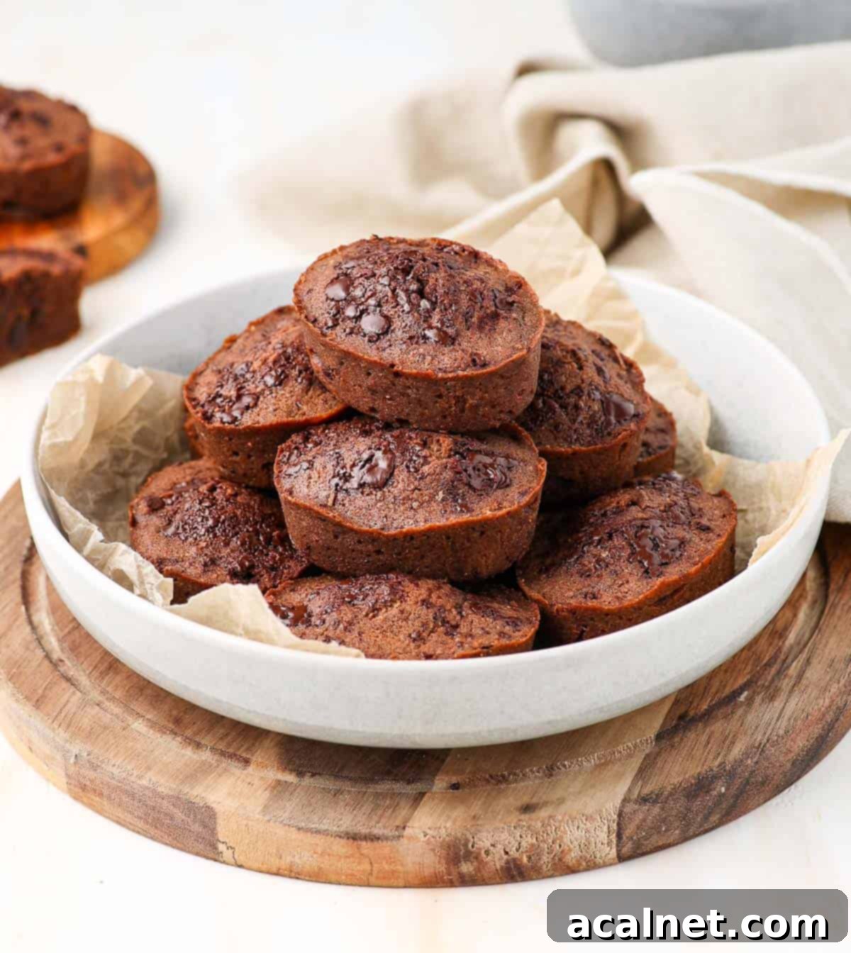 Decadent Chocolate Financiers 6 Several chocolate financiers stacked neatly in a large white bowl, placed on a round wooden cutting board, invitingly ready to be enjoyed.