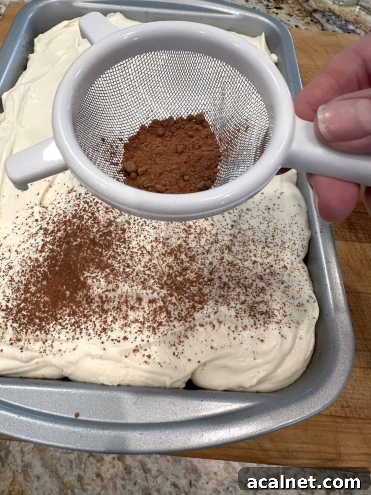 A small fine-mesh strainer dusting cocoa powder over the mascarpone-frosted tiramisu cake.