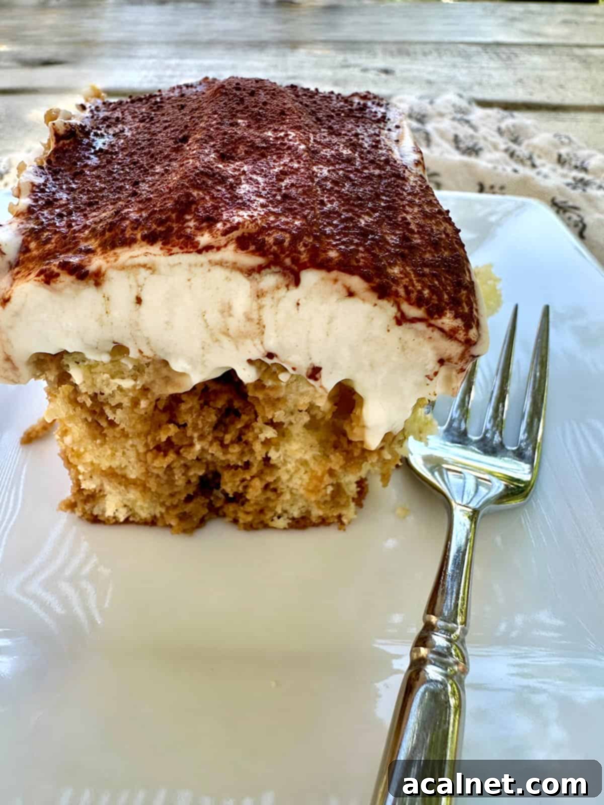 A perfectly portioned serving of tiramisu cake on a white plate with a fork, showing the distinct layers of cake and creamy frosting.