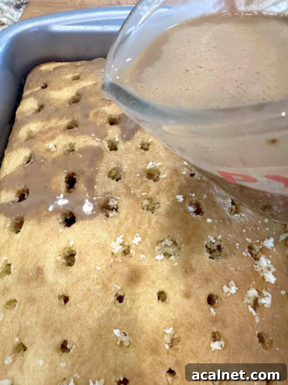 A measuring cup full of coffee mixture being poured slowly over a vanilla cake with small holes poked into it.