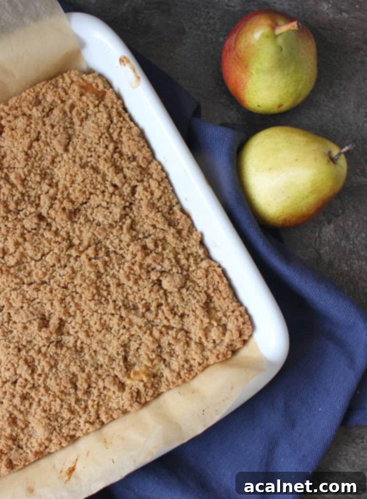 Overhead view of a baked Pear Crumble Cake next to two fresh pears, emphasizing the golden streusel topping and inviting texture.