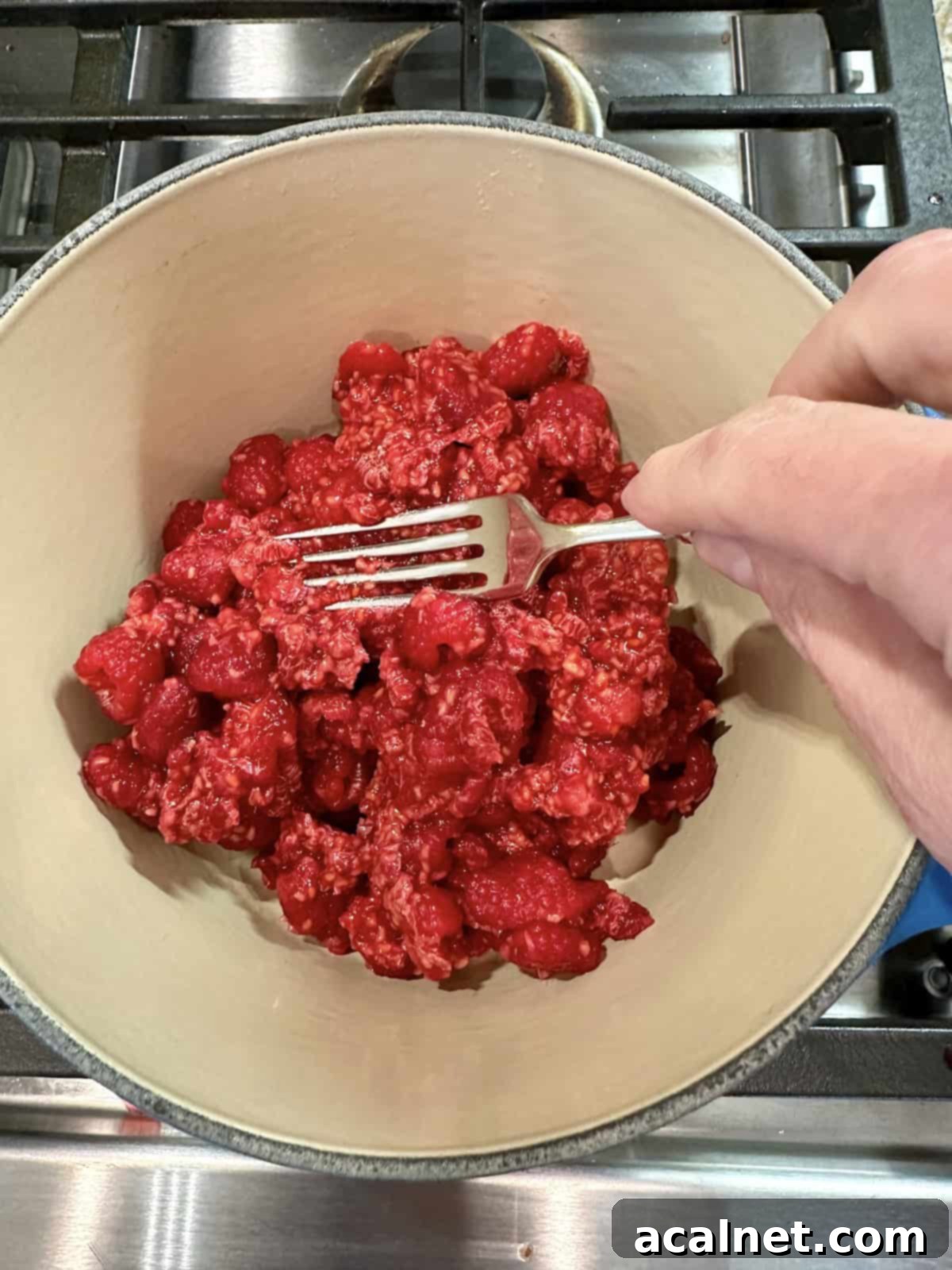A fork mashing fresh raspberries in a pan on the stove.