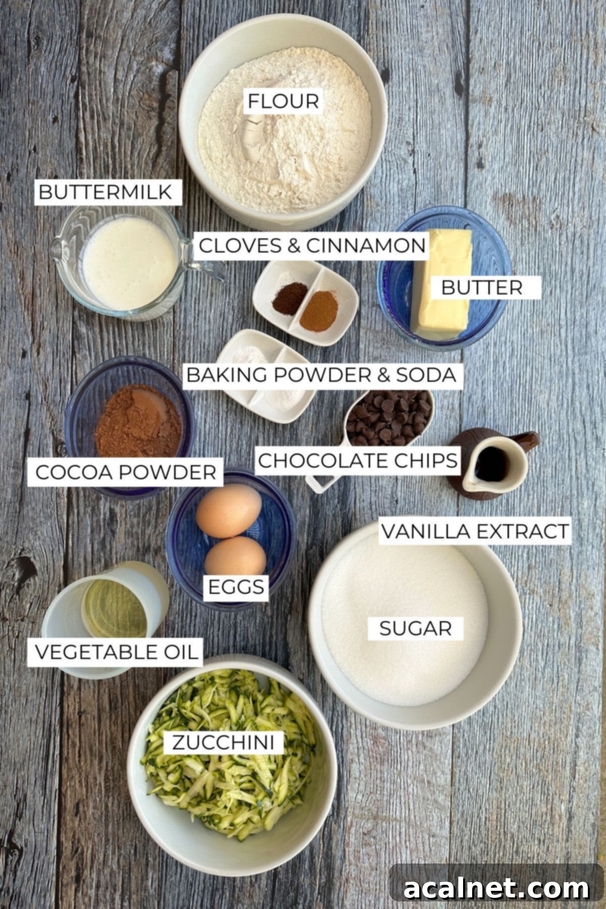 A collection of ingredients for chocolate zucchini cake, including fresh zucchini, cocoa powder, and spices, laid out on a wooden table.