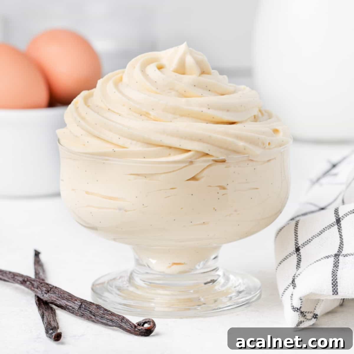 A glass cup filled with perfectly piped Vanilla Bavarian Cream, elegantly garnished and surrounded by fresh vanilla beans, eggs, and milk, highlighting the key ingredients.