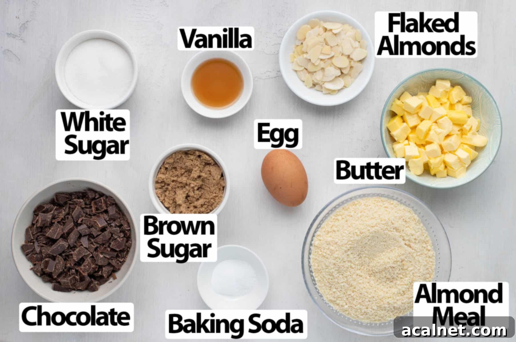 A selection of cookie ingredients including butter, sugars, egg, almond meal, and chocolate chips, neatly arranged over a white surface.