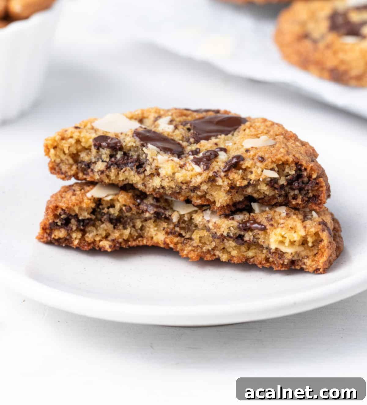 A chocolate chip almond cookie cut in half, stacked artfully on a small white plate, showing its soft interior.