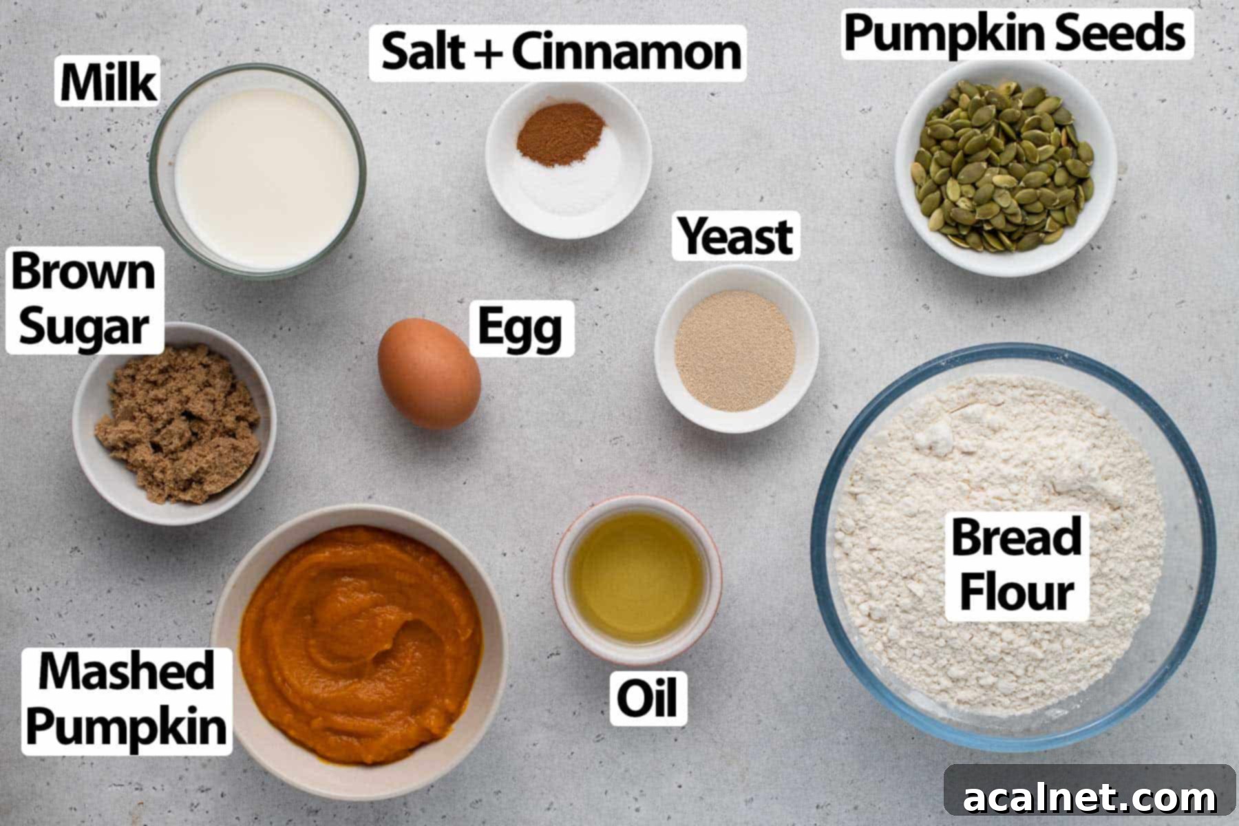 A flat lay of various ingredients for pumpkin buns, including flour, pumpkin puree, milk, eggs, and spices, arranged on a light grey surface.