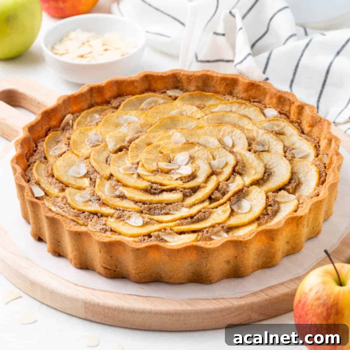 Elegant Apple Frangipane Tart 2 A perfectly baked Apple Frangipane Tart on a rustic wooden board, surrounded by fresh red apples and a striped tea towel, showcasing its golden crust and artfully arranged apple slices.