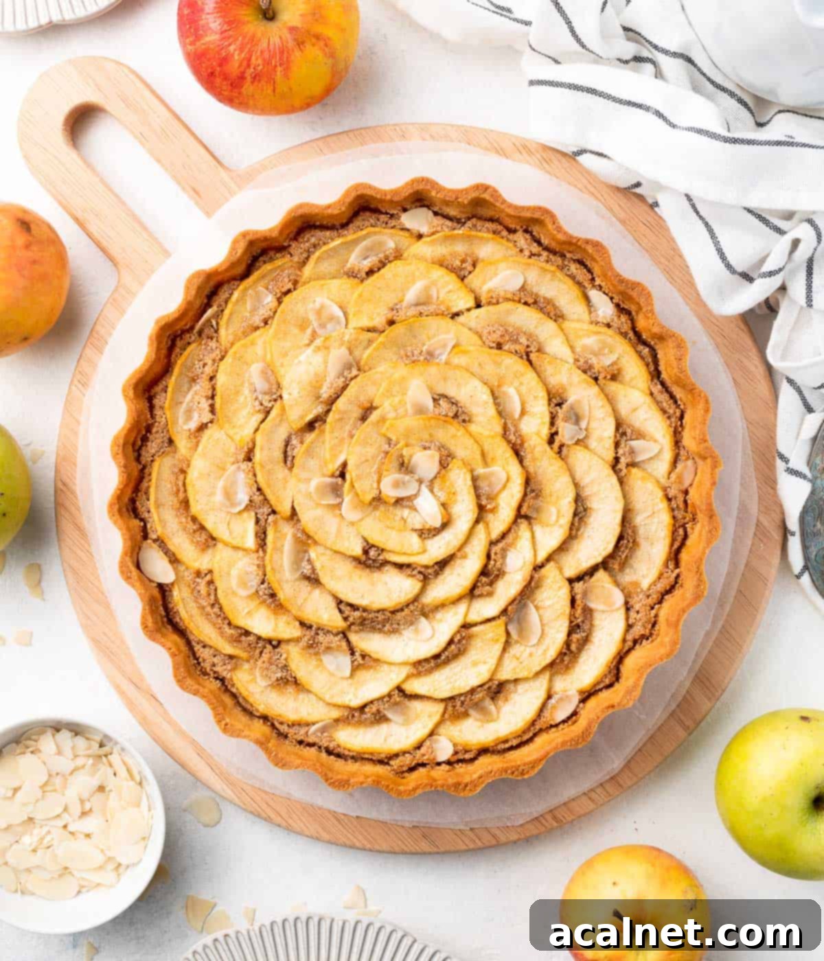 Elegant Apple Frangipane Tart 8 A top-down view of a golden-brown Apple Frangipane Tart, artfully arranged with apple slices and a hint of almond cream, resting on a wooden serving board on a white surface.