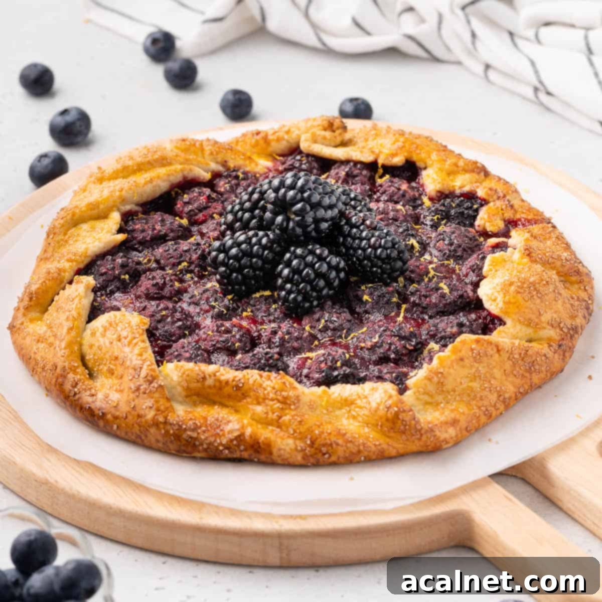 A freshly baked rustic blackberry tart, garnished with fresh blackberries, resting on a sheet of baking paper over a round wooden board. Its golden, flaky crust perfectly encases the juicy, dark berry filling.