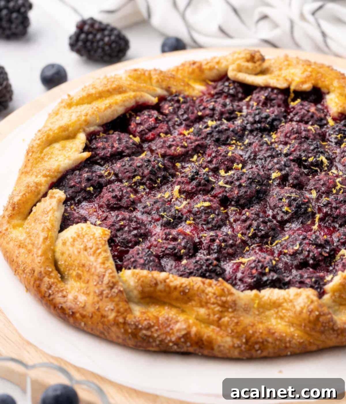 A beautifully baked blackberry galette, viewed from the side, resting on a round wooden board with some fresh blackberries scattered around.