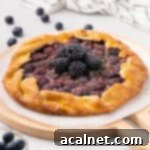 A freshly baked rustic blackberry tart, garnished with fresh blackberries, resting on a sheet of baking paper over a round wooden board. Its golden, flaky crust perfectly encases the juicy, dark berry filling.