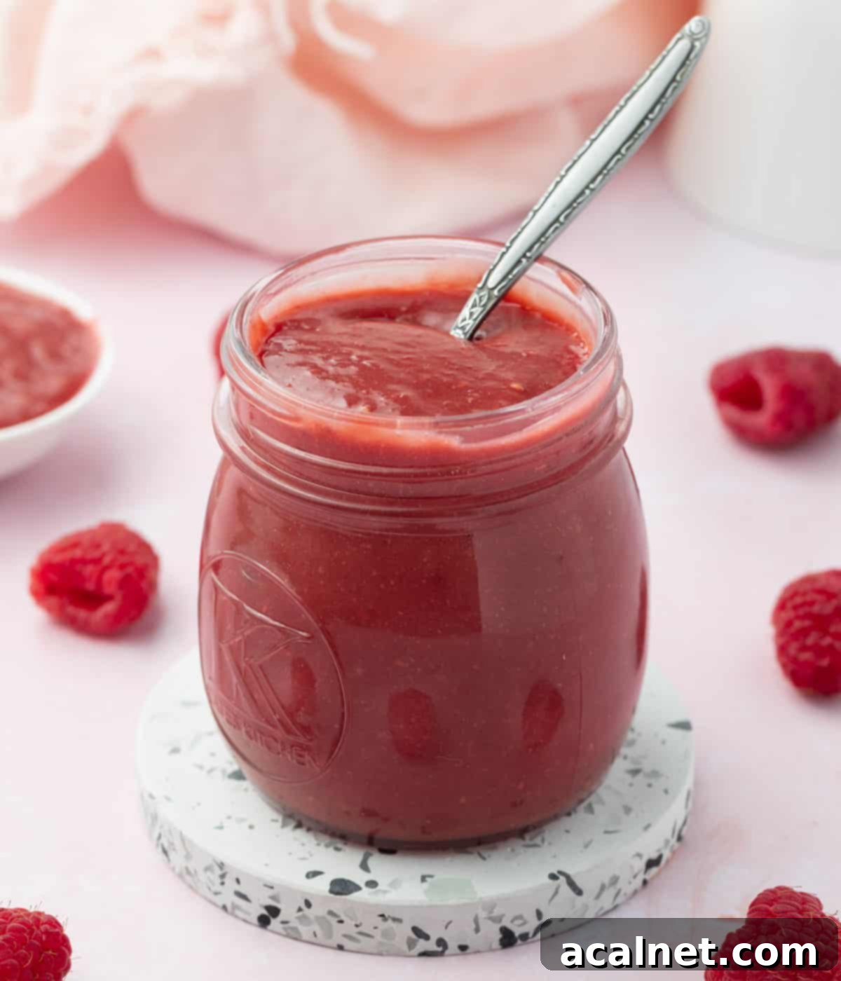 Set raspberry curd inside a glass jar with a silver spoon on a soft pink background.