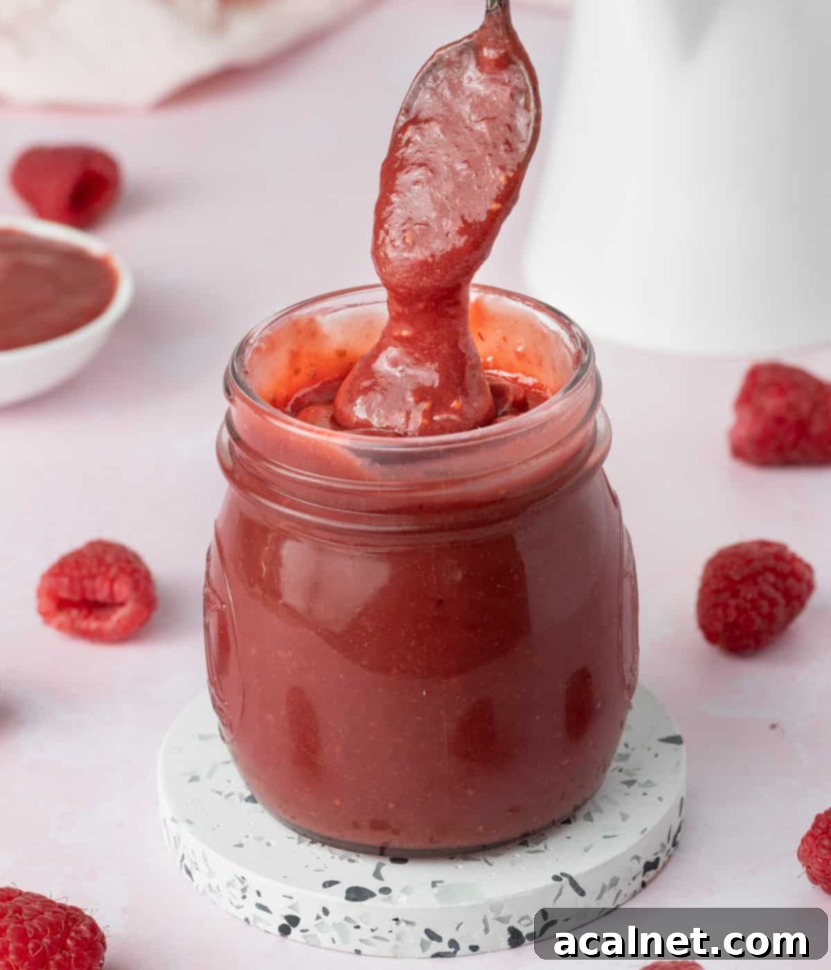 Creamy raspberry curd dripping from a silver spoon back into a glass jar, showcasing its smooth texture.