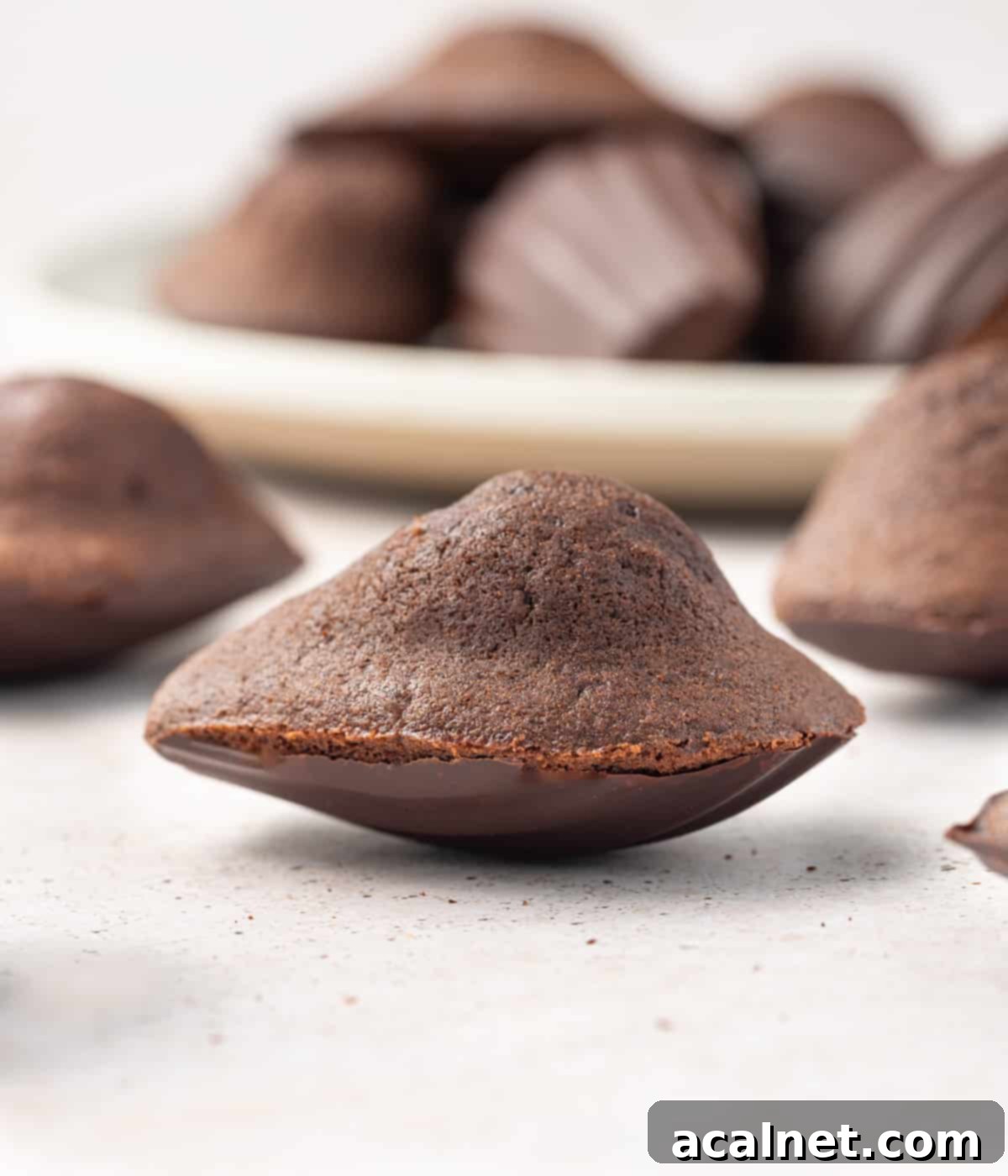 close up on one madeleine on a beige surface with a bowl of madeleines in the background.