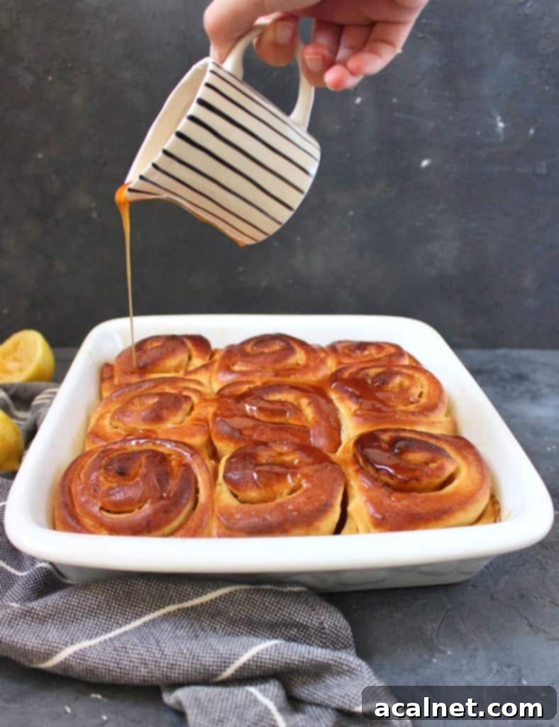 A hand pouring glossy lemon caramel over freshly baked Lemon Sweet Rolls in a white baking dish.