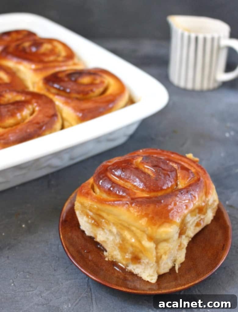 A single Lemon Sweet Roll on a small brown plate, with the baking dish of other rolls blurred in the background.