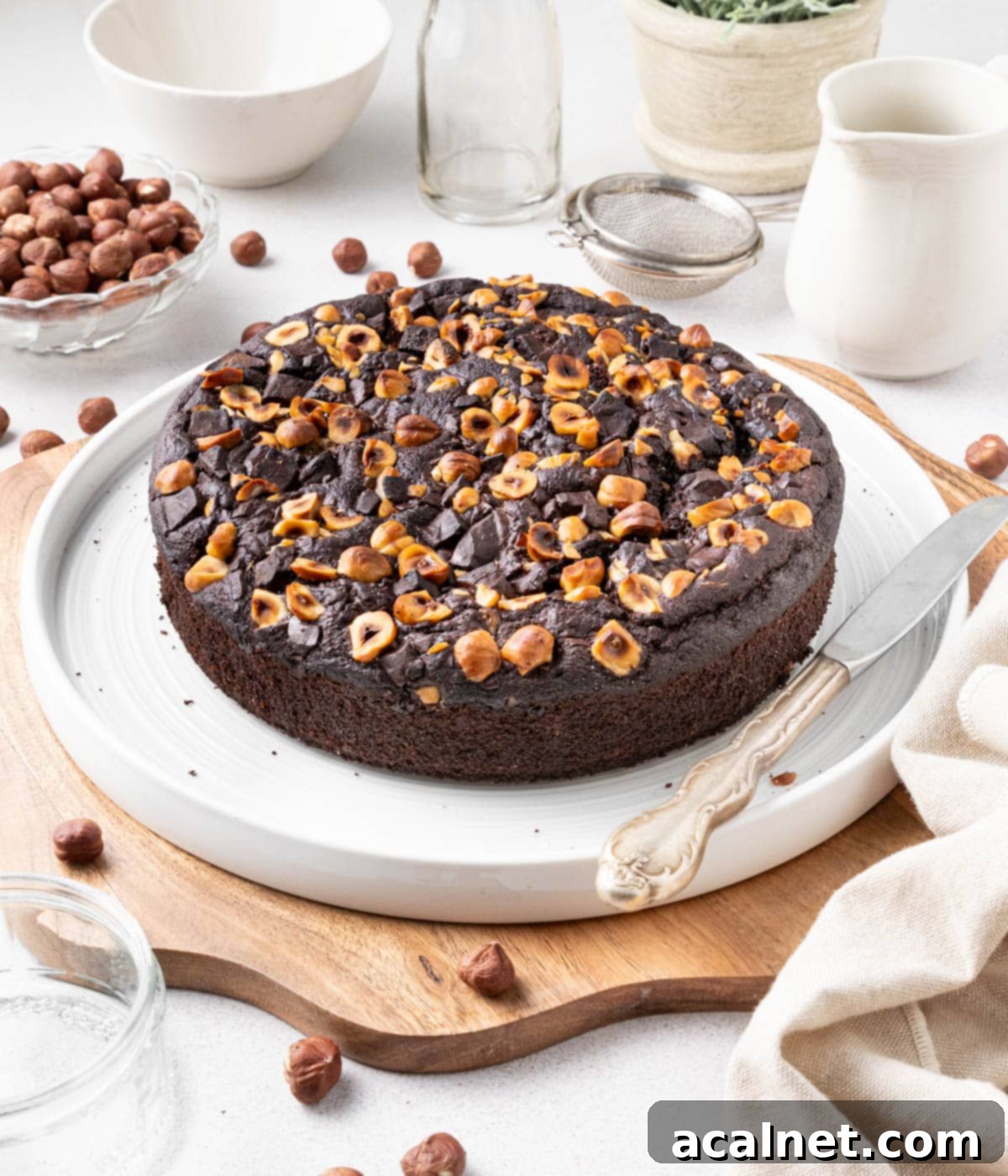 Simple Chocolate Hazelnut Delight 8 A whole Chocolate Hazelnut Cake presented on a large white plate, nestled on a wooden cutting board, surrounded by elegant crockery and a soft beige tea towel, setting the scene for a delightful afternoon treat.