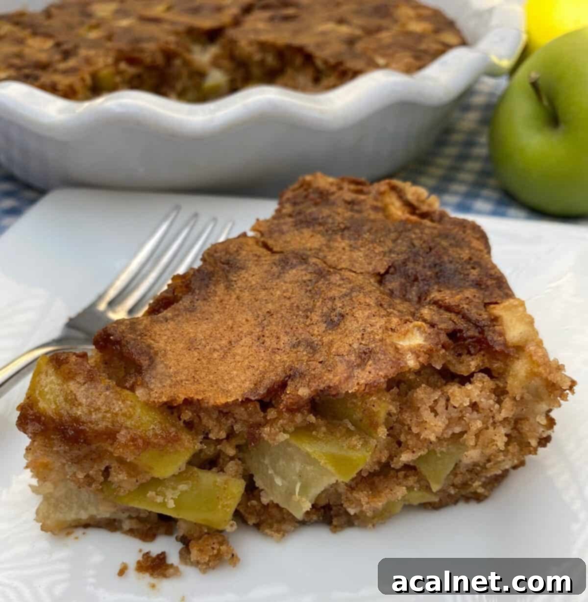 A wedge of apple cake with the baking dish and an apple in the background.