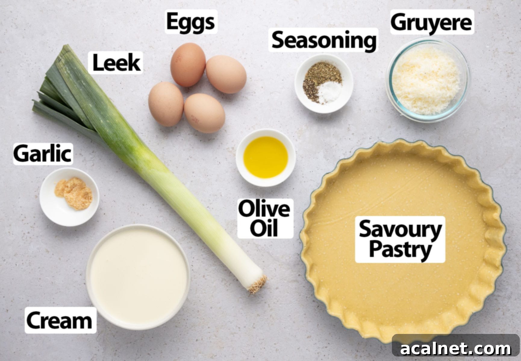 Ingredients laid on a light grey surface, including flour, butter, leeks, eggs, cream, and a block of Gruyère cheese.