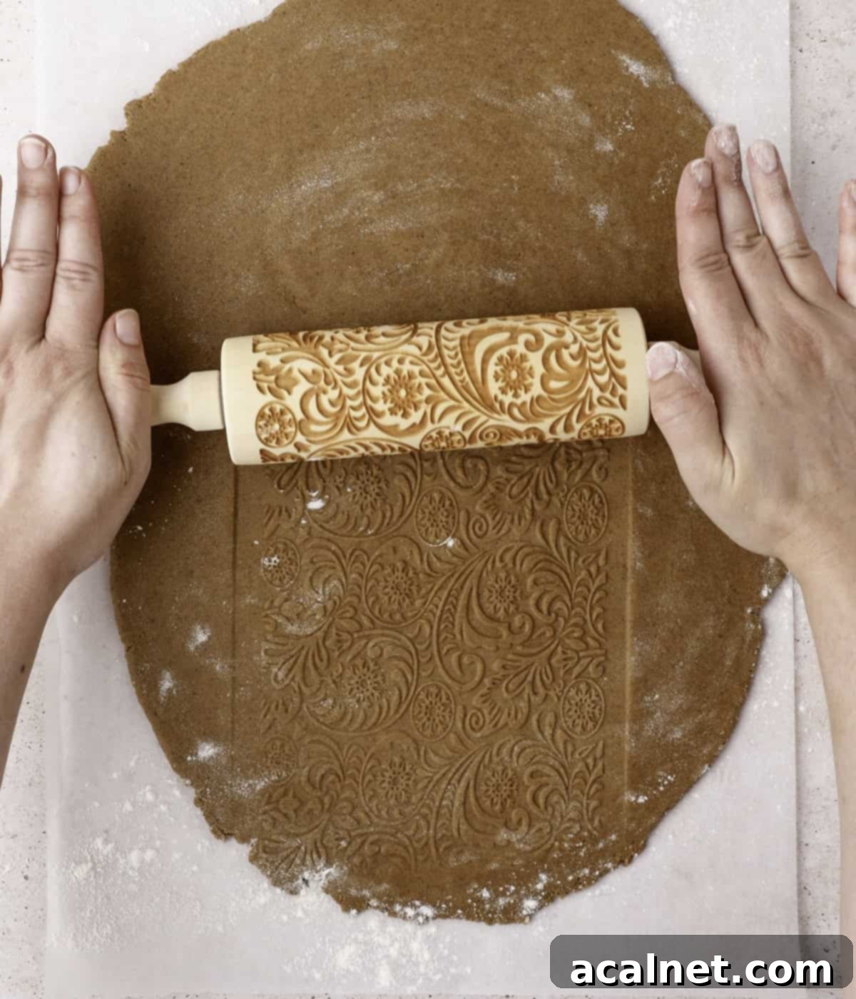 An embossed rolling pin being carefully rolled over flattened cookie dough to create a decorative pattern, ready for cutting.