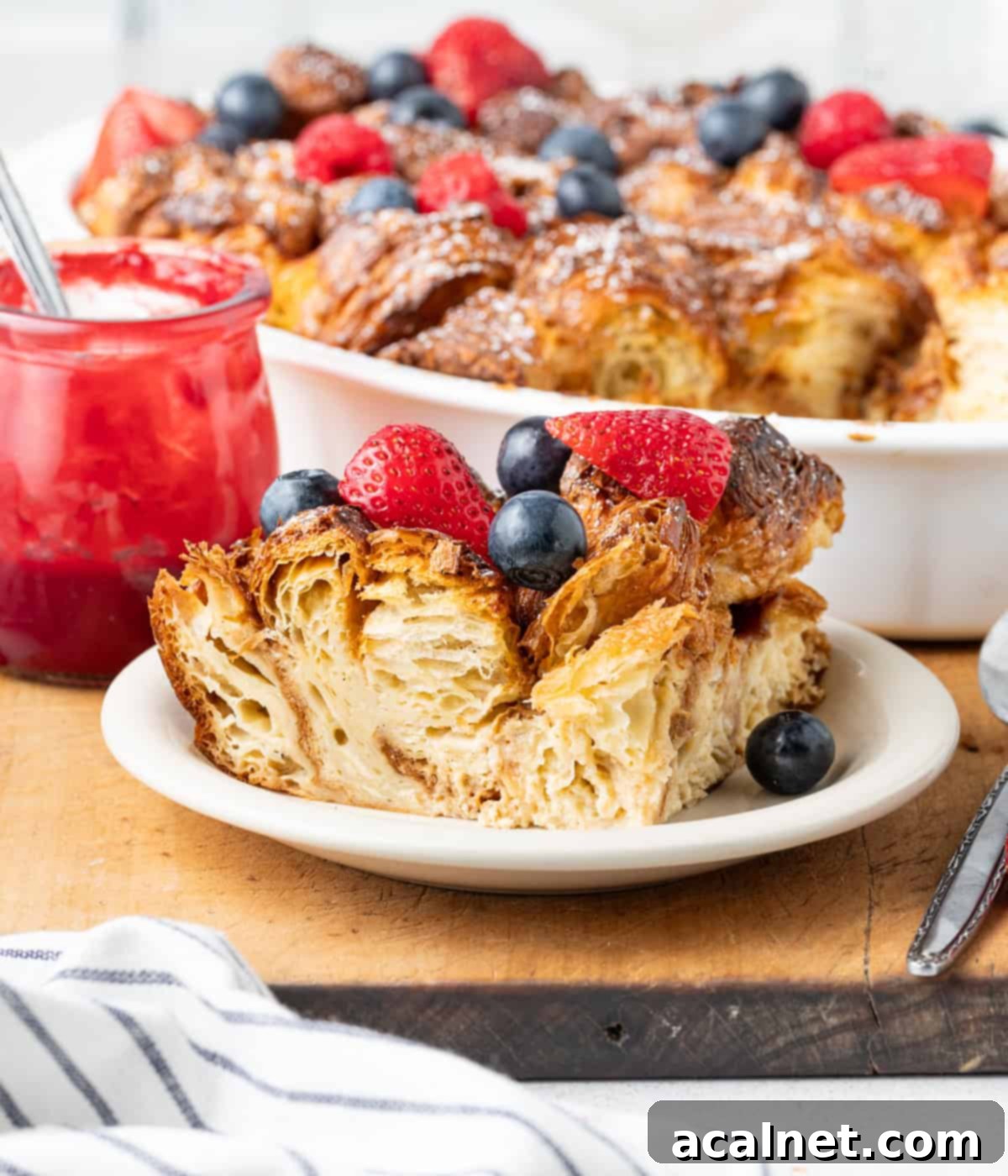 Slice of french toast bake on a small white plate topped with berries, with the rest of the dish in the background.