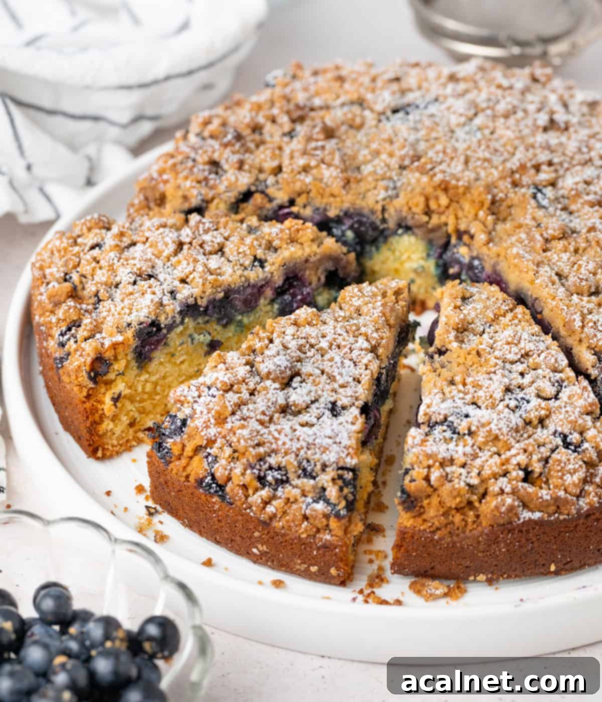 A whole Blueberry Sour Cream Coffee Cake on a large white plate with three slices already cut out, ready to be served.