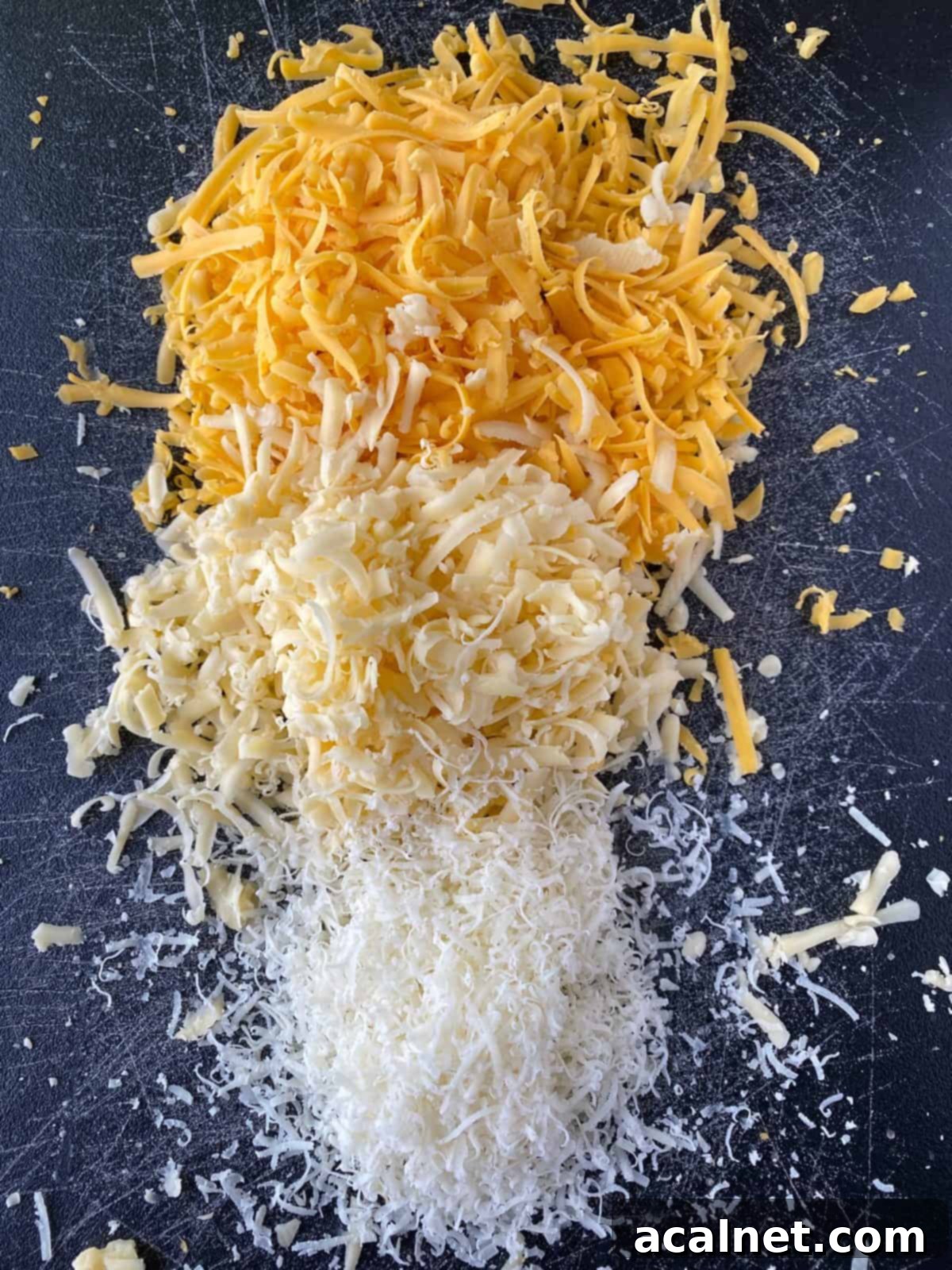 Three distinct mounds of freshly grated cheese – cheddar, smoked gouda, and parmesan – arranged on a wooden cutting board.