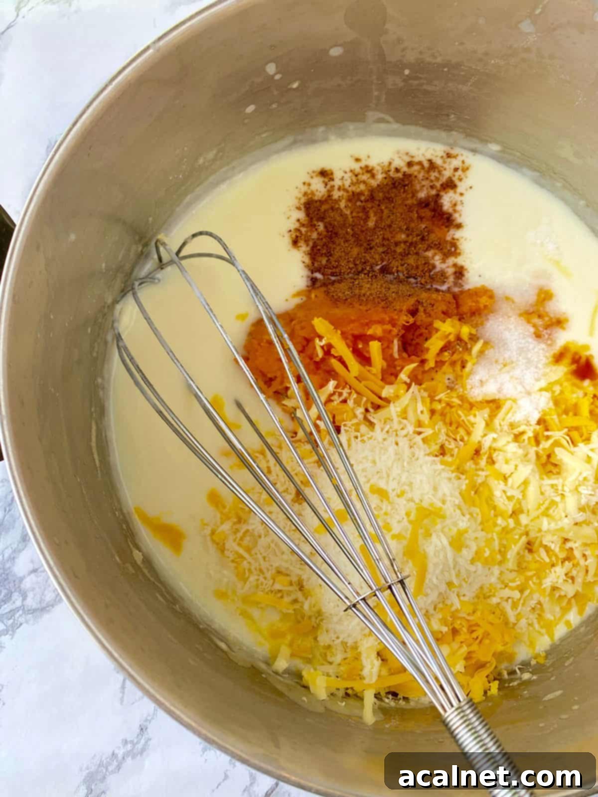 A saucepan on the stove containing a creamy white sauce being whisked, with piles of shredded cheeses, cooked butternut squash, and spices nearby, ready to be incorporated.