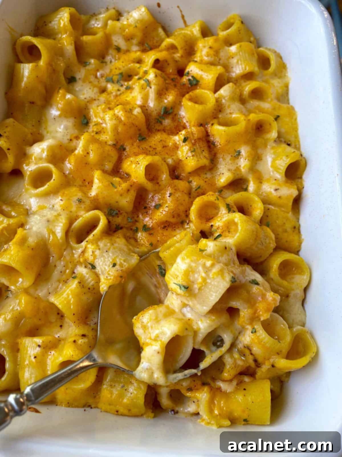 A large ceramic baking dish of golden butternut squash mac and cheese, with a serving spoon taking a portion from the bubbly, browned surface.