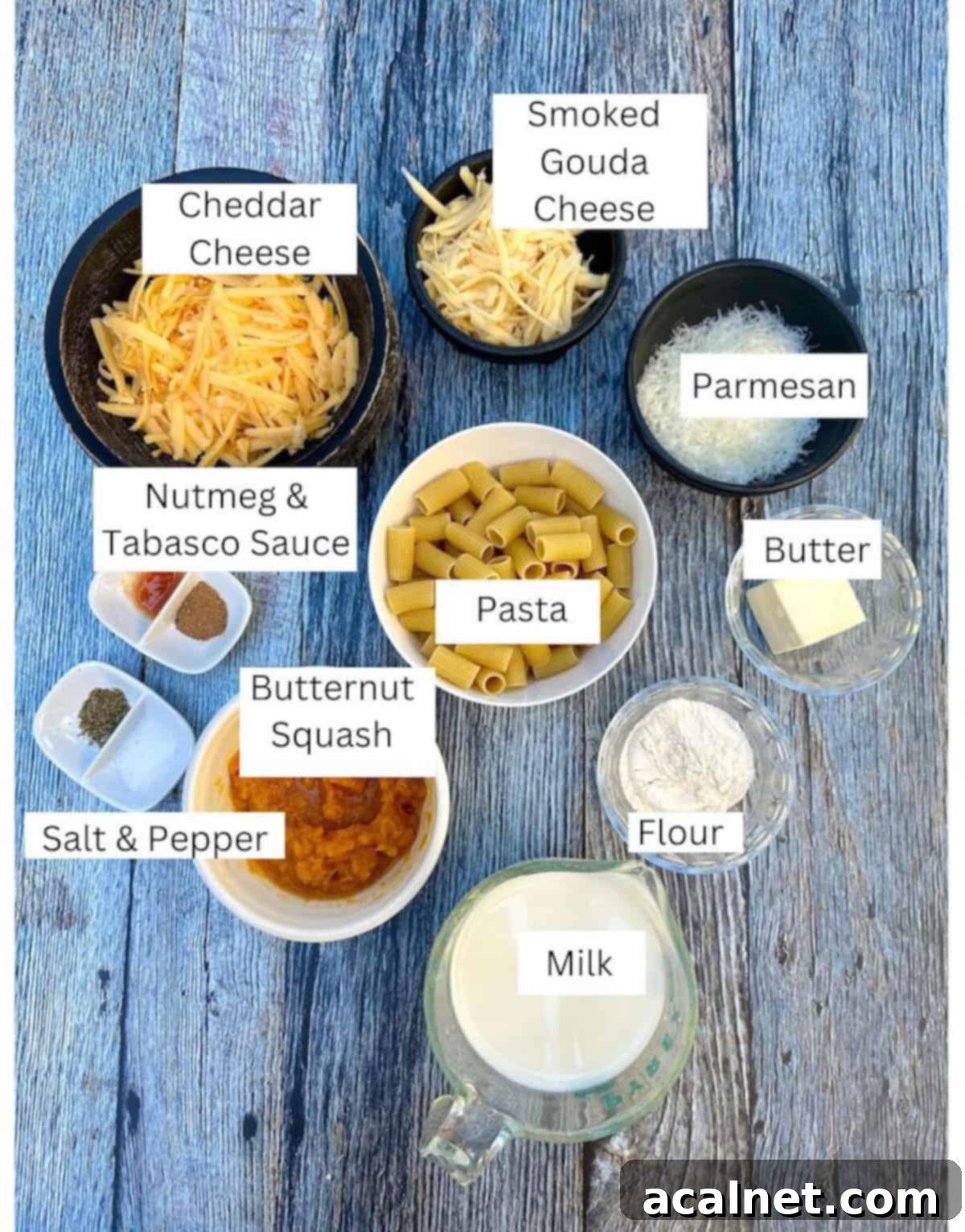 A collection of ingredients for Butternut Squash Mac and Cheese, neatly arranged in small white dishes on a wooden board. Labels indicate each ingredient, including shredded cheeses, diced butternut squash, pasta, and spices.