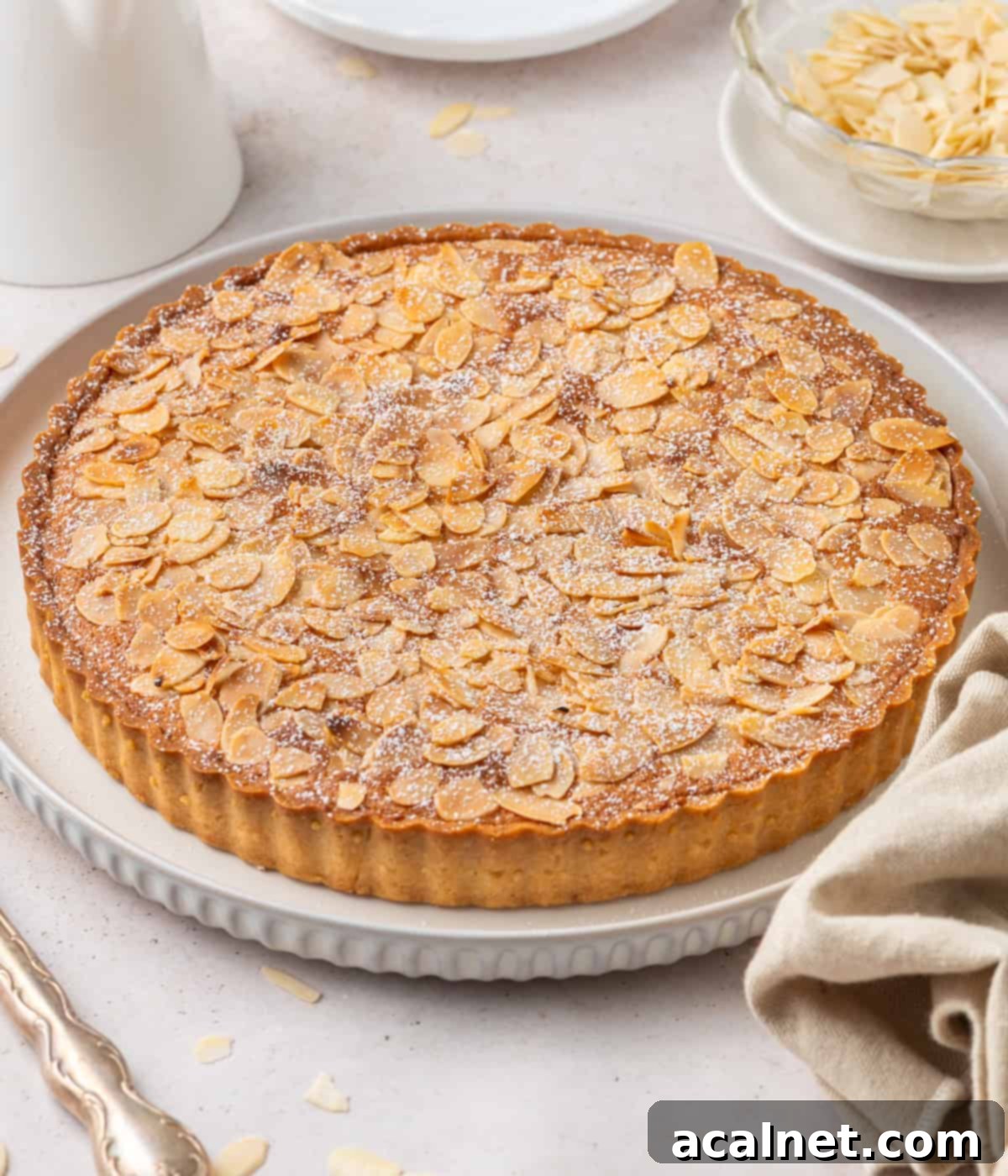 A beautifully baked French Almond Tart, whole and inviting, resting on a white serving plate over a light beige surface, ready to be enjoyed.