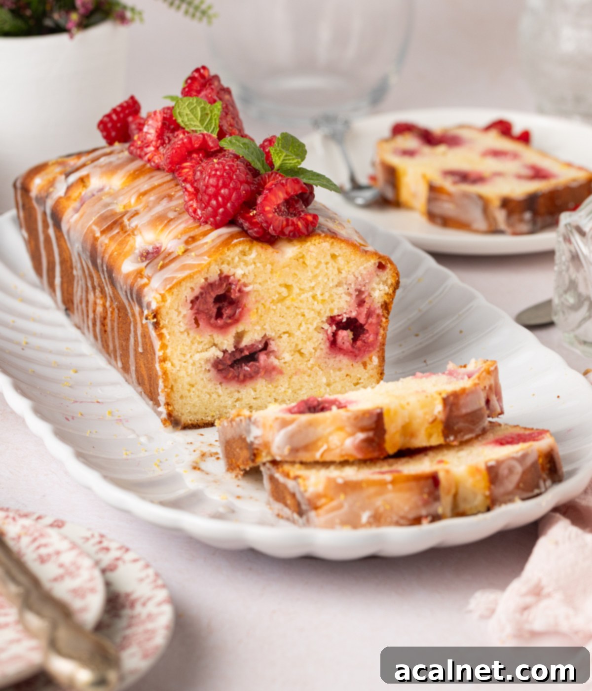 Zesty Lemon Raspberry Loaf 6 Loaf with two slices of cake placed on a white oval plate, and a slice of cake on small plate in the background.