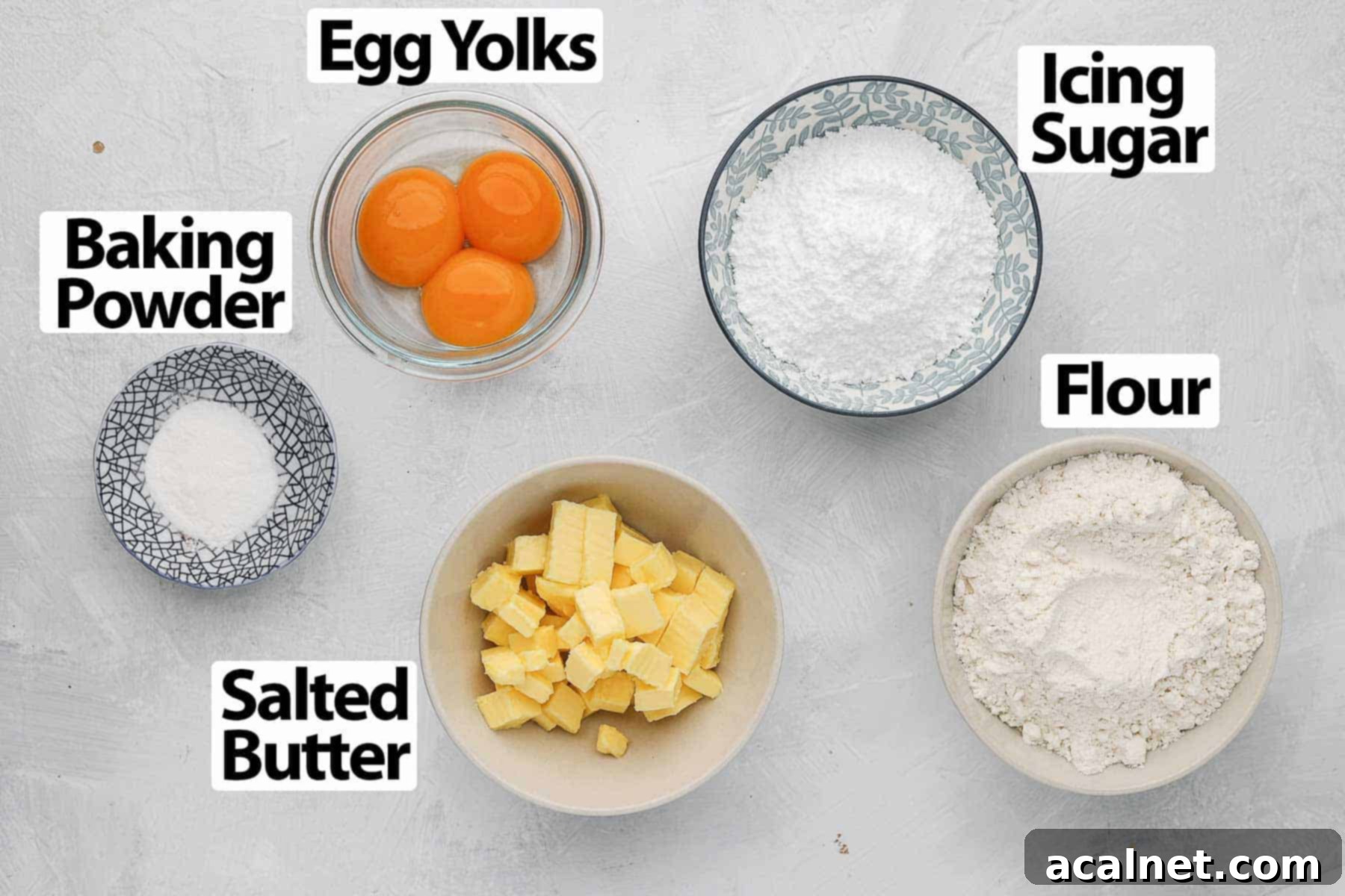 Buttery Breton Cookie Delights 4 All 5 ingredients for Sablés Bretons Cookies laid out on a white surface: egg yolks, icing sugar, salted butter, flour, and baking powder.