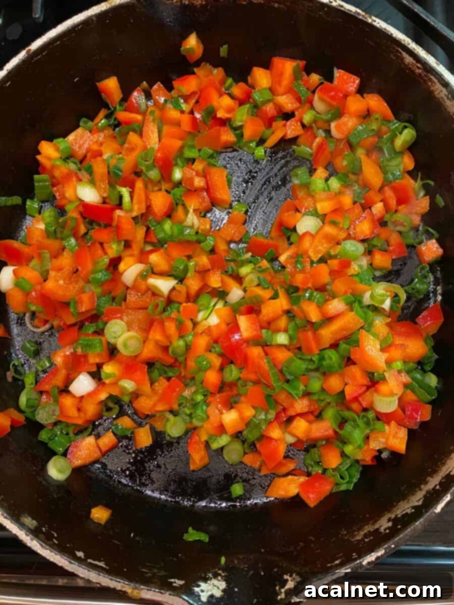 Diced red peppers and chopped green onions sizzling in a skillet on the stove.