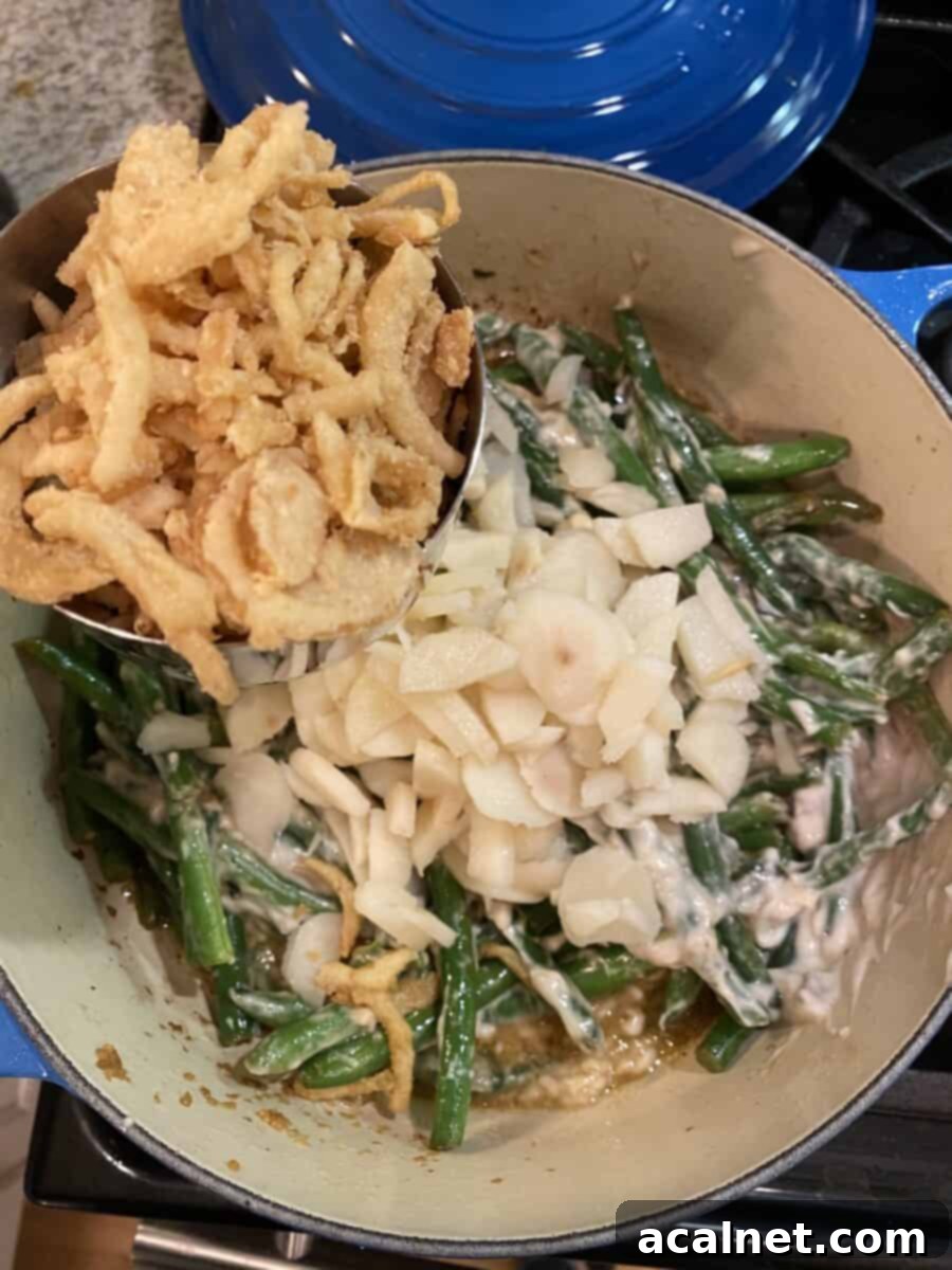 A pan on the stovetop filled with sautéed green beans, diced water chestnuts, and a portion of crispy fried onions, stirred together before baking.