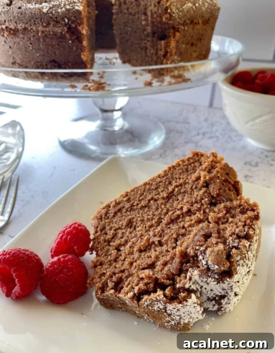 A slice of cake with raspberries with the entire cake on a serving plate in the background.