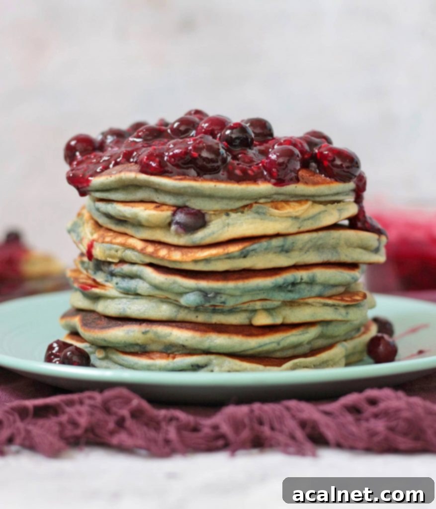 A stack of golden brown blueberry pancakes generously drizzled with mixed berry compote, placed on a green plate with a purple tea towel in the background.