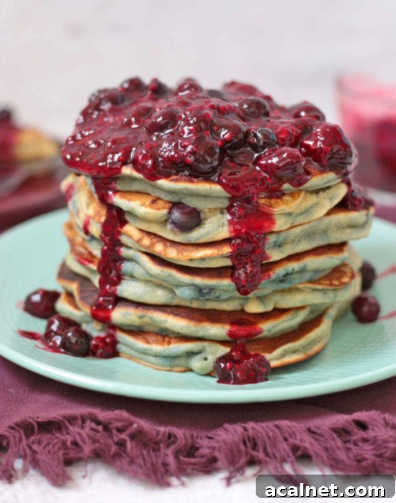 Close-up of a stack of fluffy blueberry pancakes with rich blueberry sauce generously dripping down the sides, garnished with fresh blueberries.