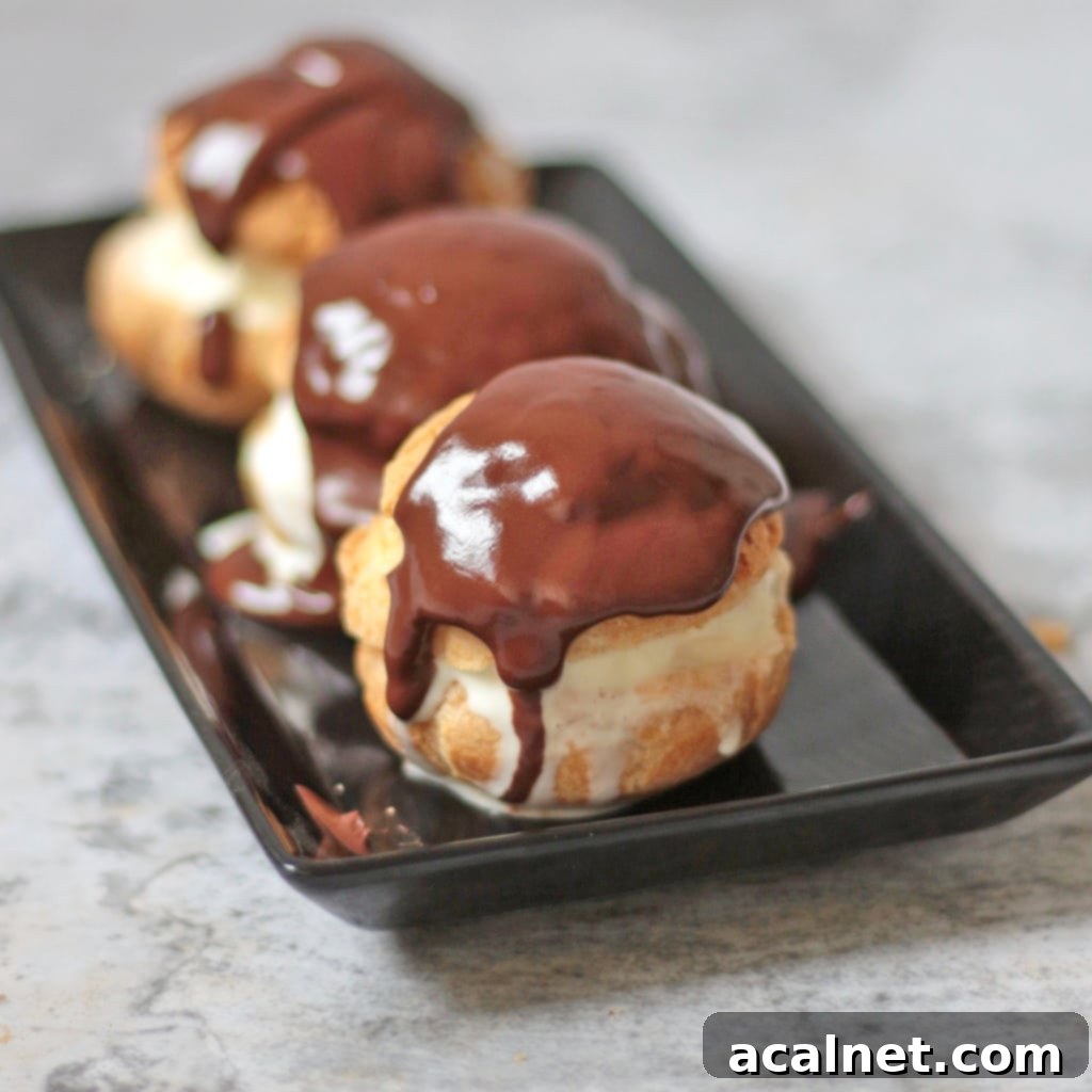 Three classic French profiteroles filled with vanilla ice cream and topped with warm chocolate sauce on a black plate.