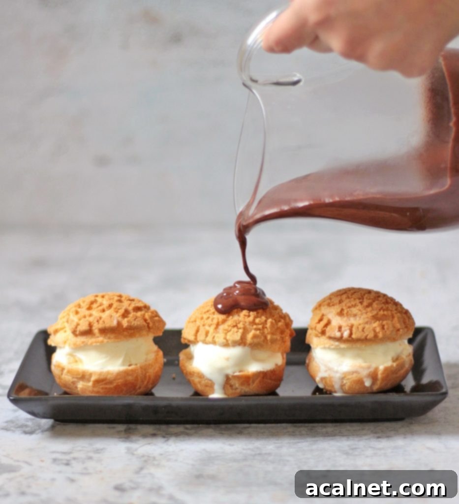 Warm chocolate sauce being drizzled over a stack of ice cream-filled profiteroles, creating a decadent dessert.